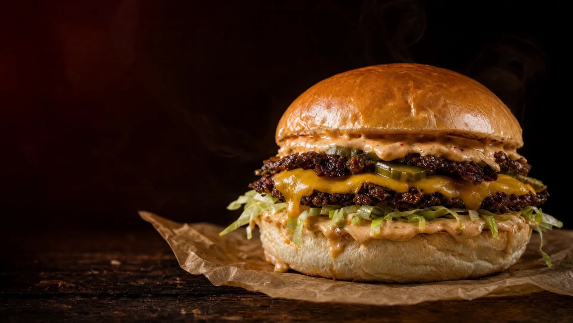 Double smash burger cu cheddar topit, iceberg, castraveți murați și sos, pe hârtie de copt, în lumină caldă